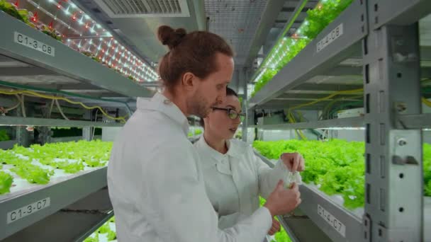 Dos científicos, un hombre y una mujer con batas blancas ponen muestras de plantas en un tubo de ensayo con el fin de averiguar los resultados de las pruebas . — Vídeos de Stock