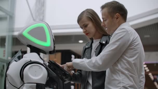 Két ember egy férfi és egy nő kommunikál a robot. Nyomja meg a robot a kezét a képernyőn. A robot asszisztens kölcsönhatásban egy bevásárlóközpontban egy pár. — Stock videók