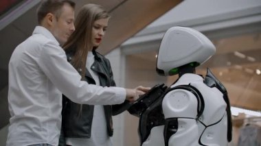 Alışveriş Merkezinde bir erkek ve bir kadın ekrana dokunarak ve gülümseyerek bir robot danışmanı ile etkileşim. Cyborg Android insanlara yardım ediyor.