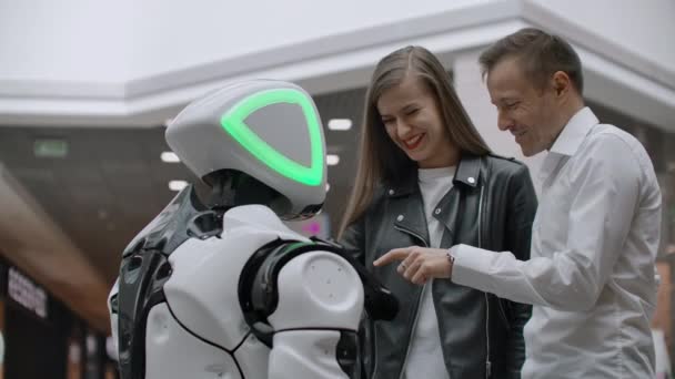 Ein Mann und eine Frau in der Mall interagieren mit einem Roboterberater, indem sie auf den Bildschirm tippen und lächeln. Cyborg-Androide helfen Menschen. — Stockvideo