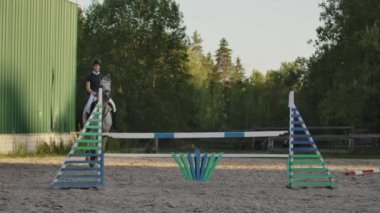 Ağır çekim, Close Up, düşük açı: Horsegirl güneşli açık havada kumlu parkour terbiye arenada çit atlama güçlü kahverengi at binme. Rekabetçi binici modacı engellerin üzerinden atlama eğitimi.