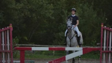 Slow Motion, Close Up: Genç atlı atlı güçlü kahverengi at abine binerek güneşli açık havada kumlu parkour binicilik arenasında çit atlama. Gün batımında engellerin üzerinden atlayan rekabetçi binici eğitimi.
