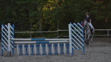 Slow Motion, Close Up: Genç atlı atlı güçlü kahverengi at abine binerek güneşli açık havada kumlu parkour binicilik arenasında çit atlama. Gün batımında engellerin üzerinden atlayan rekabetçi binici eğitimi.