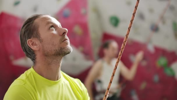 Ritratto di bellissimo uomo scalatore di roccia che assicura un altro scalatore con corda. Interni parete di arrampicata artificiale e attrezzature — Video Stock