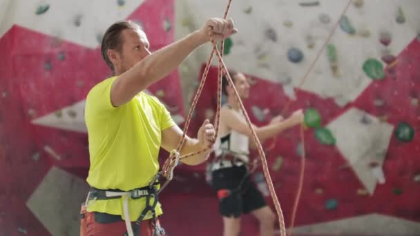 Portrait of beautiful man rock climber belaying another climber with rope. Indoors artificial climbing wall and equipment — Stock Video