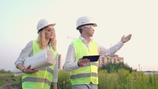 İki endüstri mühendisi güvenlik kaskı takıyor ve tablet mühendisliğini çalışıyor ve çizimlerle konuşuyor. Dışarıdaki binada. Mühendislik araçları — Stok video