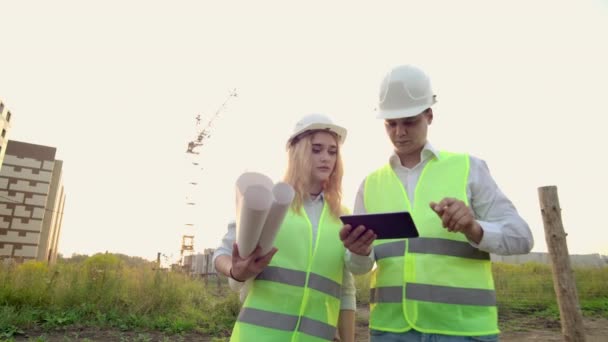 Vuxen ingenjör man och arkitekt kvinna använder en tablett i drift. Skriver ett meddelande eller kontrollerar en ritning. Mot bakgrunden bygger. — Stockvideo