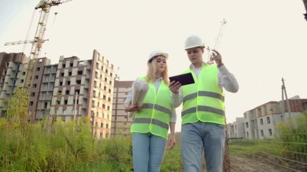 Бизнес, строительство, промышленность, технологии и люди - улыбающийся строитель в твердой шляпе с планшетным компьютером вместе с женщиной с чертежами строителей на стройке . — стоковое видео