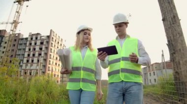 İki endüstri mühendisi güvenlik kaskı takıyor ve tablet mühendisliğini çalışıyor ve çizimlerle konuşuyor. Dışarıdaki binada. Mühendislik araçları