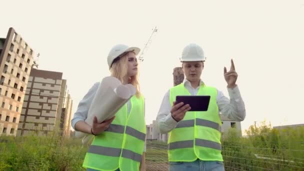 Two industrial engineer wear safety helmet and holding tablet engineering working and talking with drawings inspection. On building outside. Engineering tools — Stock Video