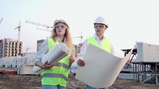 El constructor y arquitecto hombre y mujer están discutiendo el plan de construcción del moderno centro de negocios de pie cerca del sitio de construcción. Van al edificio. — Vídeos de Stock