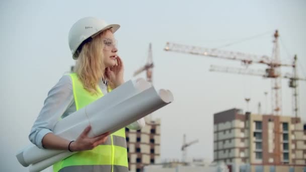 Mluvící žena v helmě na telefonu na pozadí výstavby s jeřáby, který drží v ruce kresby. Ženský inženýr na staveništi.