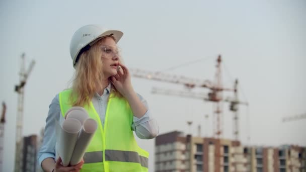 Retrato de una mujer ingeniera en un casco hablando por teléfono en el fondo de la construcción con grúas sosteniendo dibujos en sus manos. Ingeniera en obra — Vídeos de Stock