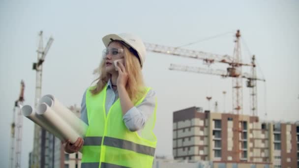 Retrato de uma mulher engenheira em um capacete falando ao telefone no fundo da construção com guindastes segurando desenhos em suas mãos. Engenheira no canteiro de obras — Vídeo de Stock