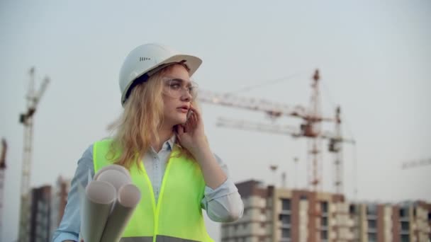 Mujer parlante en un casco en el teléfono en el fondo de la construcción con grúas con dibujos en la mano. Ingeniera en obra . — Vídeos de Stock