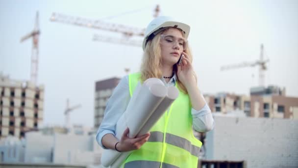 Diseñadora mujer ingeniero hablando por teléfono con el contratista con dibujos en la mano en el fondo de los edificios en construcción y grúas . — Vídeos de Stock