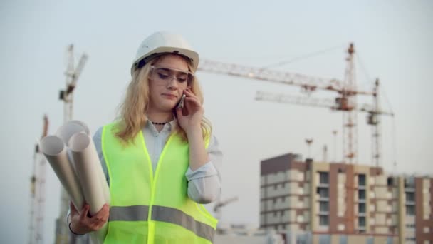 Diseñadora mujer ingeniero hablando por teléfono con el contratista con dibujos en la mano en el fondo de los edificios en construcción y grúas . — Vídeos de Stock