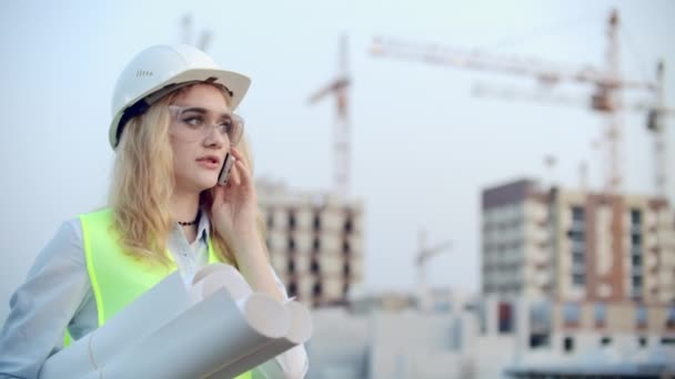 Retrato de una mujer ingeniera en un casco hablando por teléfono en el fondo de la construcción con grúas sosteniendo dibujos en sus manos. Ingeniera en obra — Vídeo de stock