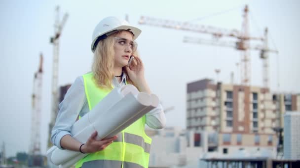 Mujer parlante en un casco en el teléfono en el fondo de la construcción con grúas con dibujos en la mano. Ingeniera en obra . — Vídeos de Stock