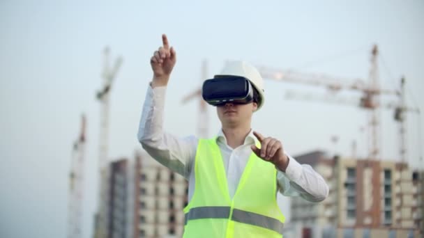 Un hombre en gafas de realidad virtual casco y chaleco en el fondo de la construcción controla las manos de la interfaz y comprueba la calidad de la construcción y el desarrollo — Vídeos de Stock