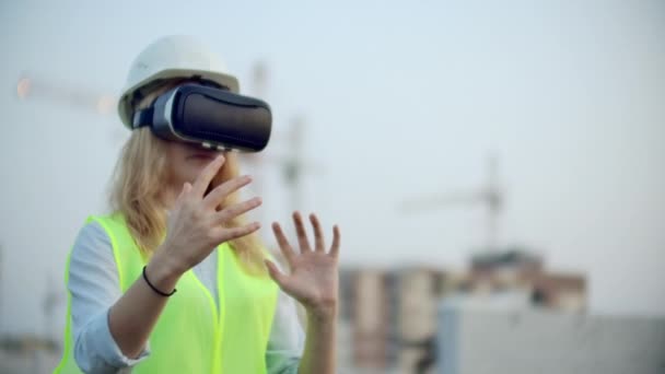 A woman in virtual reality glasses helmet and vest on the background of construction controls the hands of the interface and checks the quality of construction and development of the project and the — Stock Video