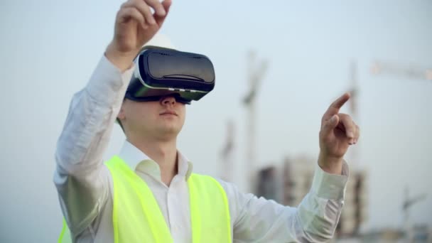 En mand i virtual reality briller hjelm og vest på baggrund af byggeriet styrer hænderne på grænsefladen og kontrollerer kvaliteten af byggeri og udvikling – Stock-video