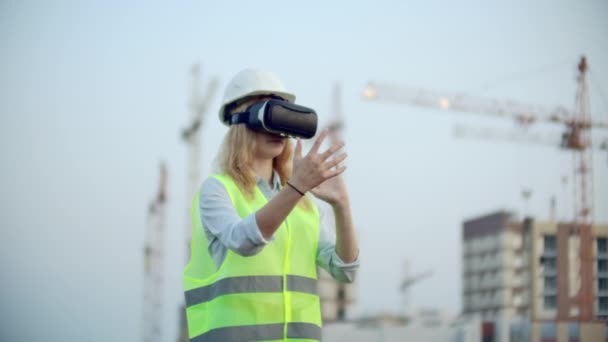 Uma mulher em óculos de realidade virtual capacete e colete no fundo da construção controla as mãos da interface e verifica a qualidade da construção e desenvolvimento do projeto e do — Vídeo de Stock