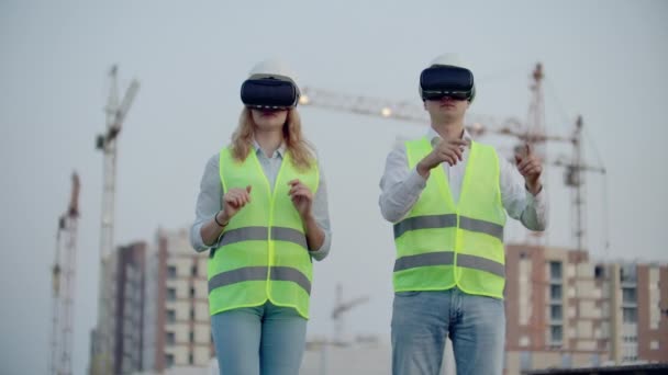 To fremtidsforvaltere på byggeplassen bruker virtuelle virkelighetsbriller på bakgrunn av bygninger og kraner, beveger hendene og klikker på ikonene – stockvideo