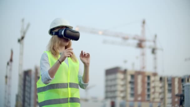 Mujer diseñadora en un sitio de construcción en casco y chaleco en las gafas de realidad virtual para mover las manos imitando la interfaz en el fondo de las grúas al atardecer — Vídeos de Stock