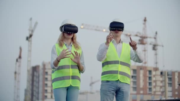 Duas pessoas Homem e mulher vestindo óculos de realidade virtual no fundo de edifícios em construção com guindastes. Gerente e assistente de design com as mãos imitando a interface — Vídeo de Stock