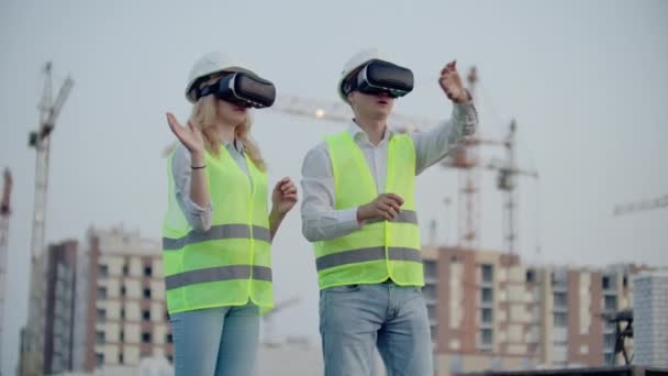 Zwei Personen in Virtual-Reality-Brillen auf dem Hintergrund von im Bau befindlichen Gebäuden mit Kränen imitieren die Arbeit der Schnittstelle zur Steuerung und Verwaltung von Bauvorhaben — Stockvideo