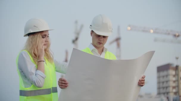 Dois construtores com desenhos em pé no fundo de edifícios em construção em capacetes e coletes, uma mulher conversando ao telefone com o cliente — Vídeo de Stock