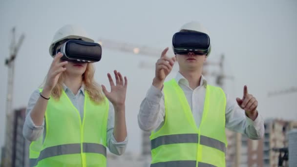 Deux personnes en lunettes de réalité virtuelle sur le fond des bâtiments en construction avec grues imitent le travail de l'interface pour le contrôle et la gestion de la construction — Video