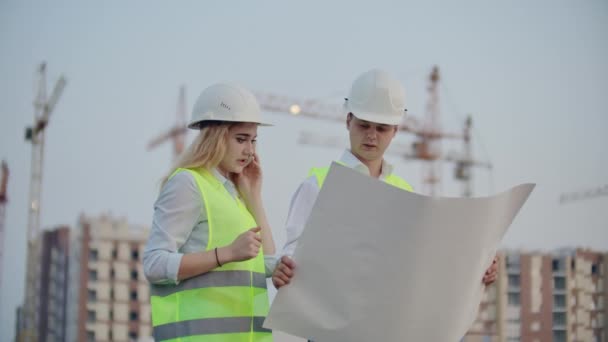 Due ingegneri discutono guardando i disegni del piano di costruzione e la posizione degli oggetti specificando l'appaltatore sui dettagli del telefono. Sto parlando al telefono al cantiere. — Video Stock