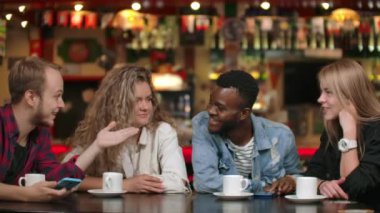 Çok uluslu erkek ve kadın grubu bir masada kahve fincanları ile birlikte oturuyor, gülümsüyor, gülüyor ve duygusal ve gesturing hikayeler anlatıyor