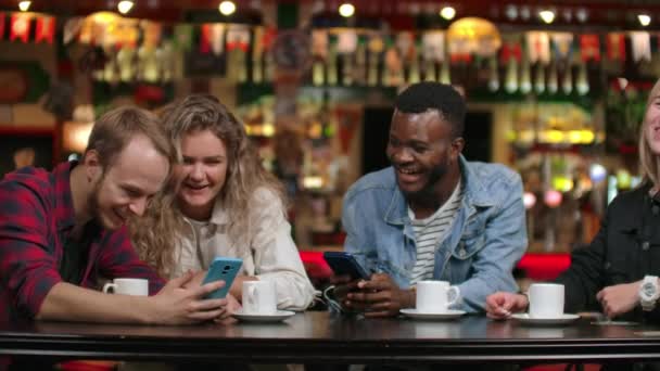 カフェに座っている4人の友人は、携帯電話の画面を見ながら笑い、旅行の写真を交換します。アフリカ系アメリカ人のガールフレンドがおしゃべりしてコーヒーを飲む. — ストック動画