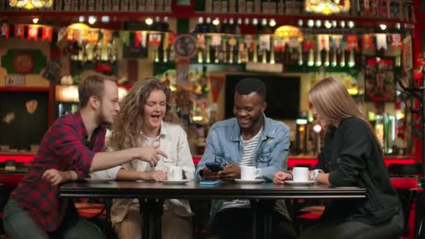 Retrato de jóvenes amigos alegres mirando el teléfono inteligente mientras están sentados en la cafetería. Gente de raza mixta sentada en una mesa en un restaurante usando teléfono móvil — Vídeos de Stock