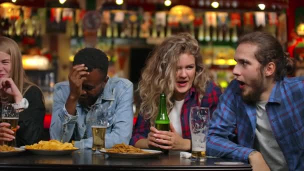 De man vertelt een grap in het gezelschap van vrienden zittend aan tafel. Meisjes en mannen lachen terwijl ze zitten aan een bar aan een tafel met bier — Stockvideo