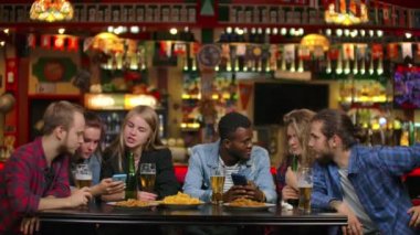 Bir adam arkadaşlarına telefonda bir fotoğraf gösterir ve herkes eşli ve bira içerken güler.