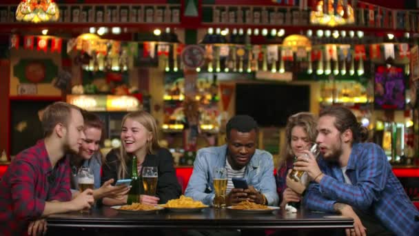 Χαρούμενοι φοιτητές που κάθονται σε ένα τραπέζι σε ένα μπαρ πίνοντας μπύρα, τρώγοντας μάρκες και παρακολουθώντας φωτογραφίες σε μια οθόνη smartphone συζητώντας φωτογραφίες — Αρχείο Βίντεο