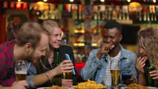 A menina mostra a seus amigos a tela de um smartphone sentado em uma mesa e todos riem enquanto estão sentados em um bar. Discuta os últimos rumores e fofocas, compartilhar fotos de viagens — Vídeo de Stock