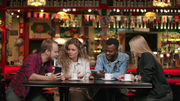 Un groupe multiethnique d'étudiants, trois Européens et un Afro-Américain, regardent les écrans téléphoniques, rient et discutent. Parlez avec des amis et regardez des photos au téléphone — Video