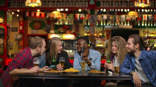Der Afroamerikaner zeigt Freunden am Telefon Bilder, sie gucken und lachen, während sie in Zeitlupe in einer Bar sitzen. Gesellschaft junger Männer und Frauen — Stockvideo
