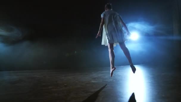 Mujer de danza moderna en un vestido blanco baila un ballet moderno, salta, hace rotación en el escenario con humo en los focos azules . — Vídeo de stock
