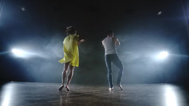 Un hombre y una mujer bailan juntos un baile divertido en jeans y un vestido amarillo en el escenario en humo. Musical — Vídeos de Stock