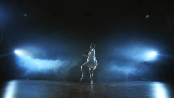 ズームカメラは、ソフトウェアと煙でステージを横切って女性ダンサーを移動します。現代の劇的なバレエ、白いドレスを着た女性が片足で回転し、ジャンプ. — ストック動画
