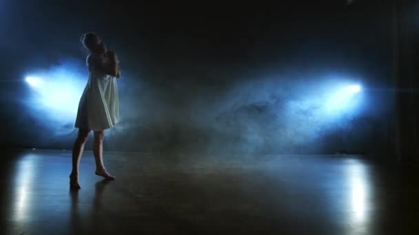 Moderní taneční žena v bílých šatech tančí moderní balet, skáče, provádí rotaci na jevišti kouřem v modrých bodových světlech. — Stock video