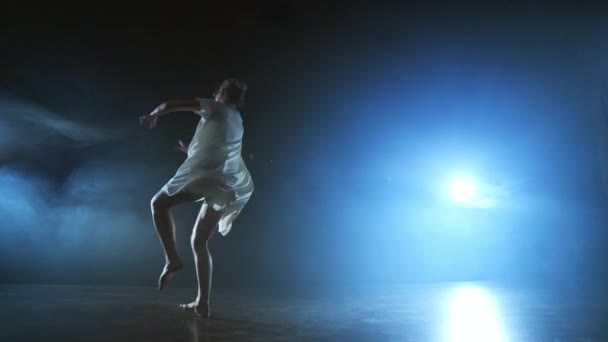 Сучасна дівчина-танцівниця в білій короткій сукні виконує пластикові красиві драматичні танці, бігаючи по сцені, падаючи на підлогу і кружляючи. Ліхтарі і дим — стокове відео