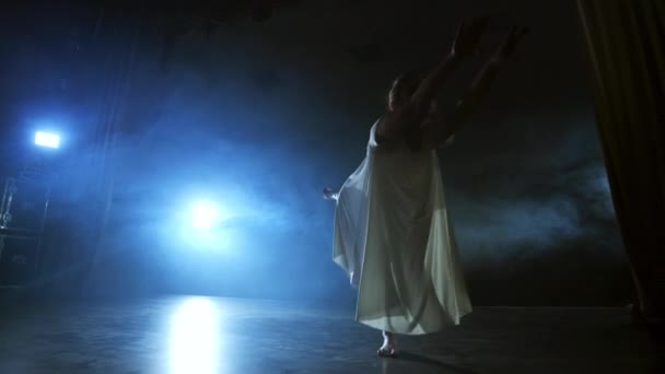 Mulher de dança moderna em um vestido branco dança um balé moderno, salta, faz rotação no palco com fumaça nos holofotes azuis . — Vídeo de Stock