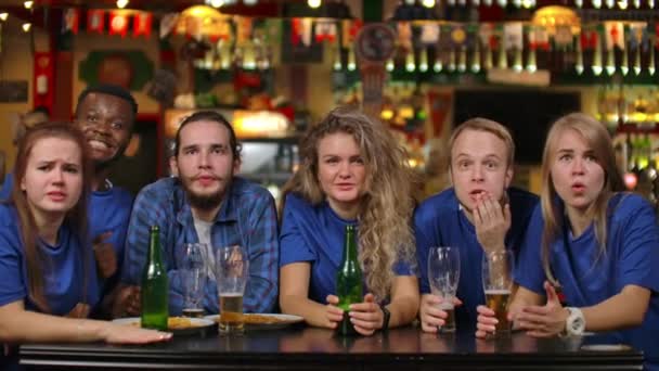 A group of young people watching the broadcast of a sporting event on TV in a beer bar, emotionally waiting for a decisive moment and feeling sad after losing a team — Stock Video